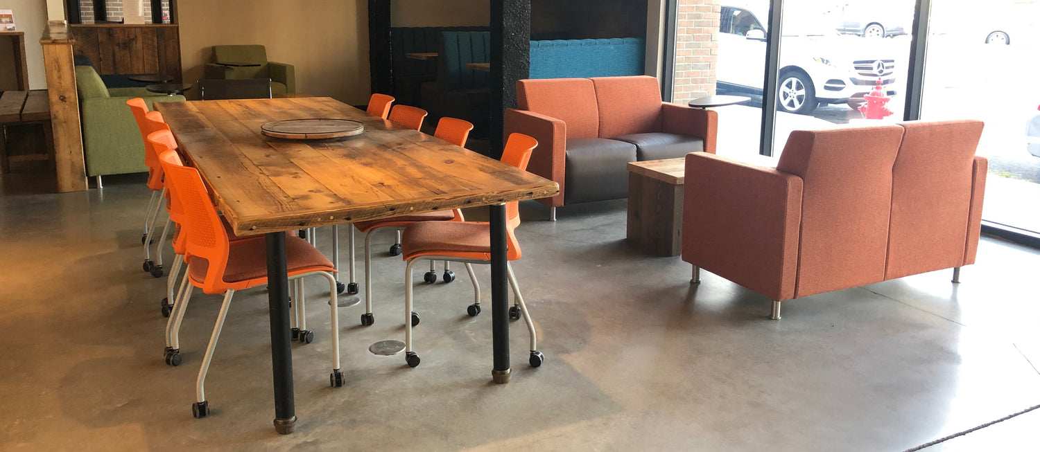 Large wooden table in the middle of the office space with eight orange rolling chairs tucked underneath. Two orange and gray softs are next to the table.